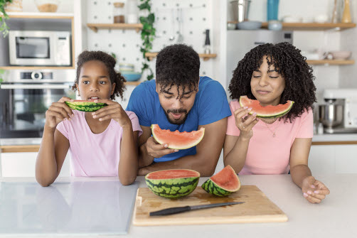 En vuxen och två barn äter vattenmelon vid köksbänken.