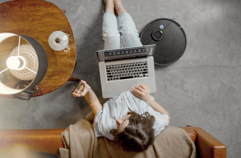 En person sitter med en laptop i knät och håller i en mobiltelefon. På golvet står en robotdammsugare.