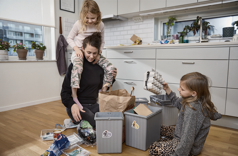 En vuxen och två barn sitter i ett kök och sorterar sopor i olika kärl