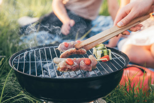 En grill med kött, korv och grönsaker står på gräset.