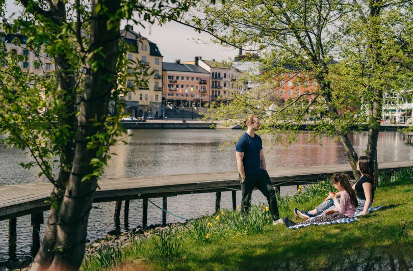 Tre personer sitter i gräset vid vattnet i Södertälje.